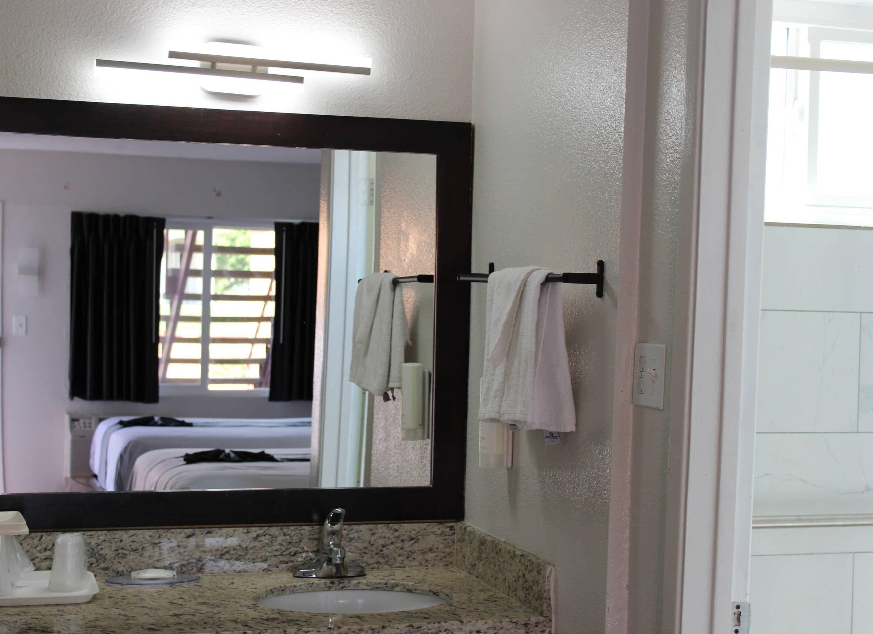 A hotel bathroom with a large mirror, a sink, a towel rack, and a bed visible in the reflection.