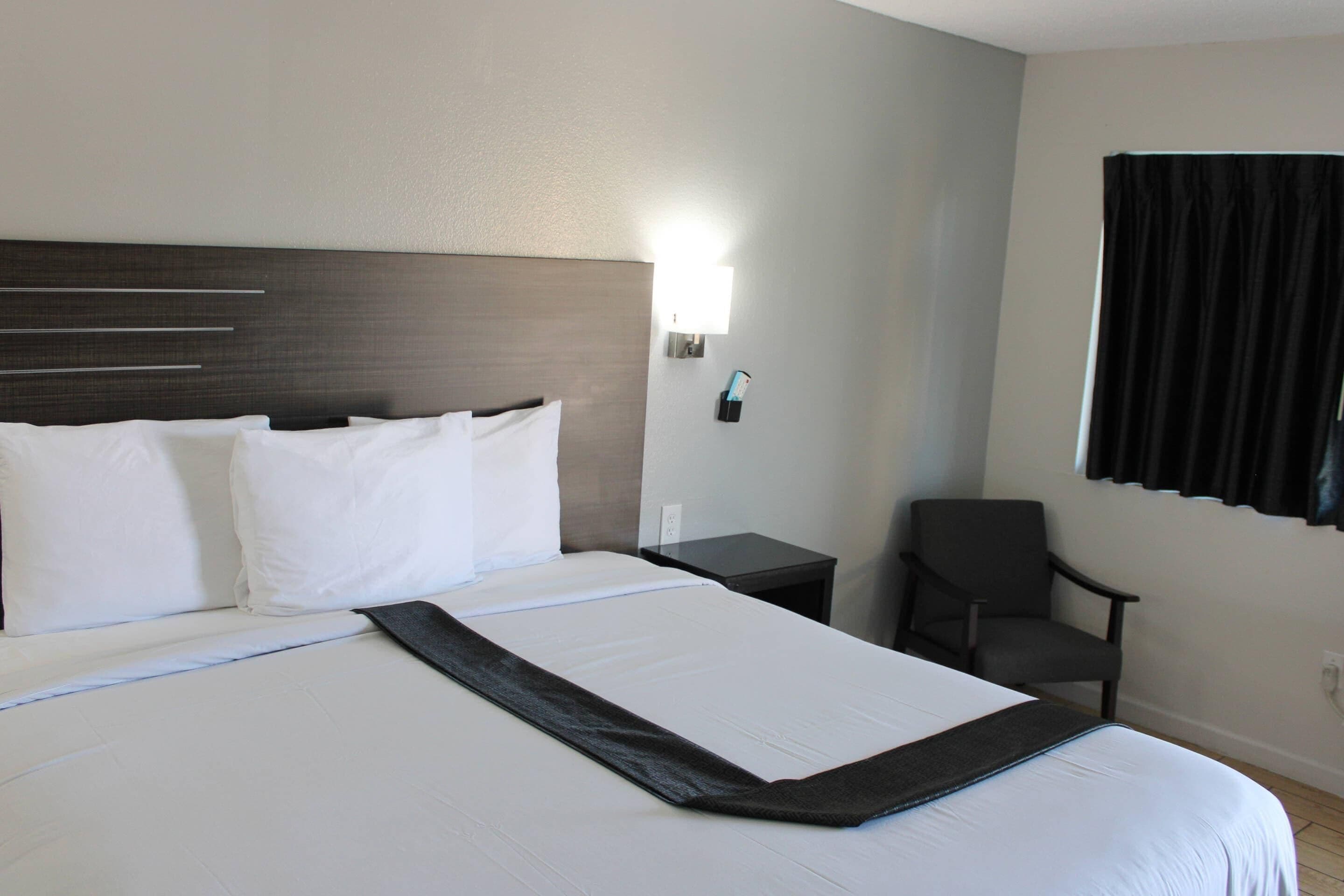 A neatly made bed with white linens and a dark accent runner, a bedside table with a lamp, and a chair in a hotel room.