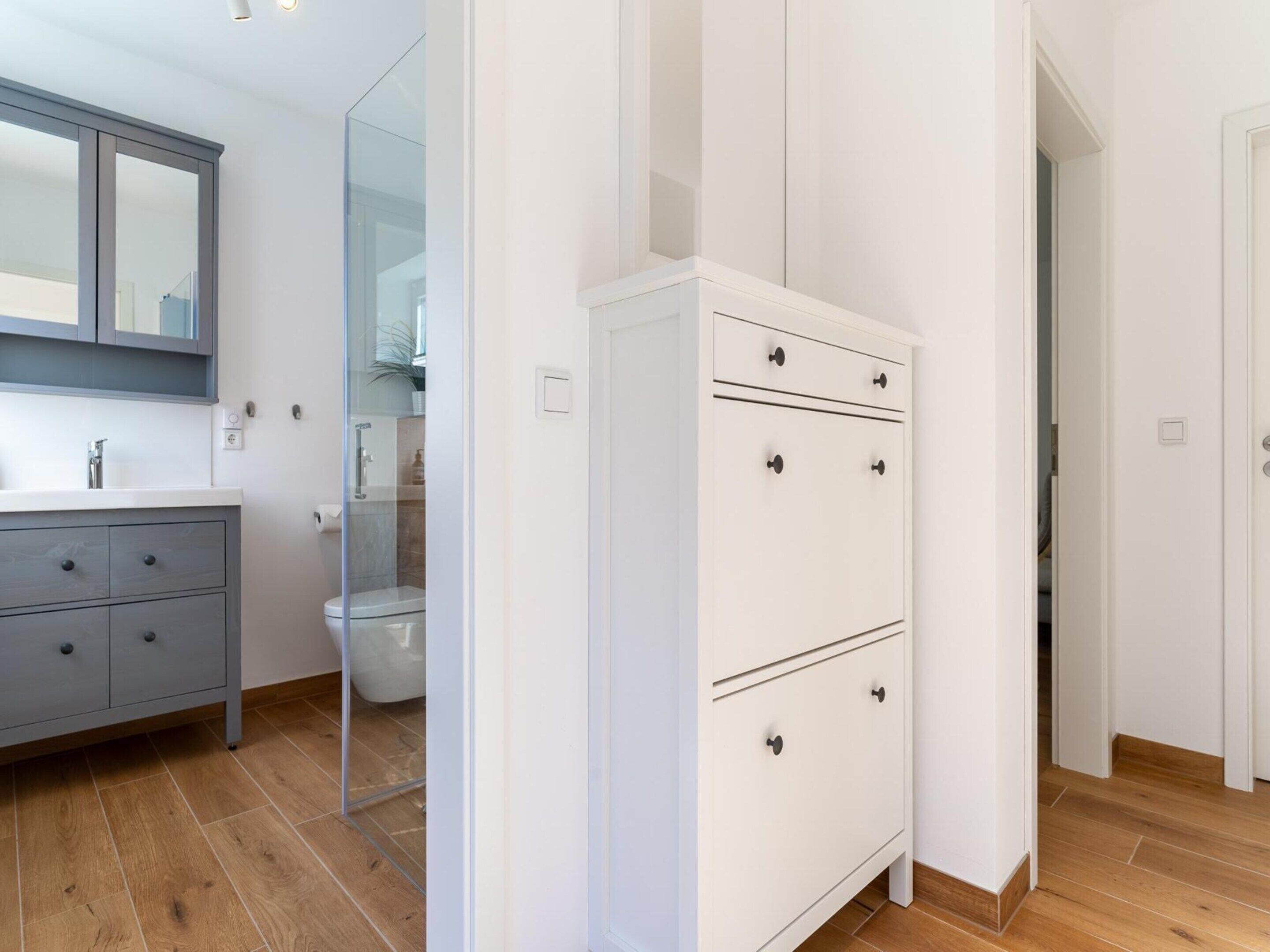 Un baño moderno con un mueble de tocador gris, una cabina de ducha de vidrio y un armario blanco con cajones.