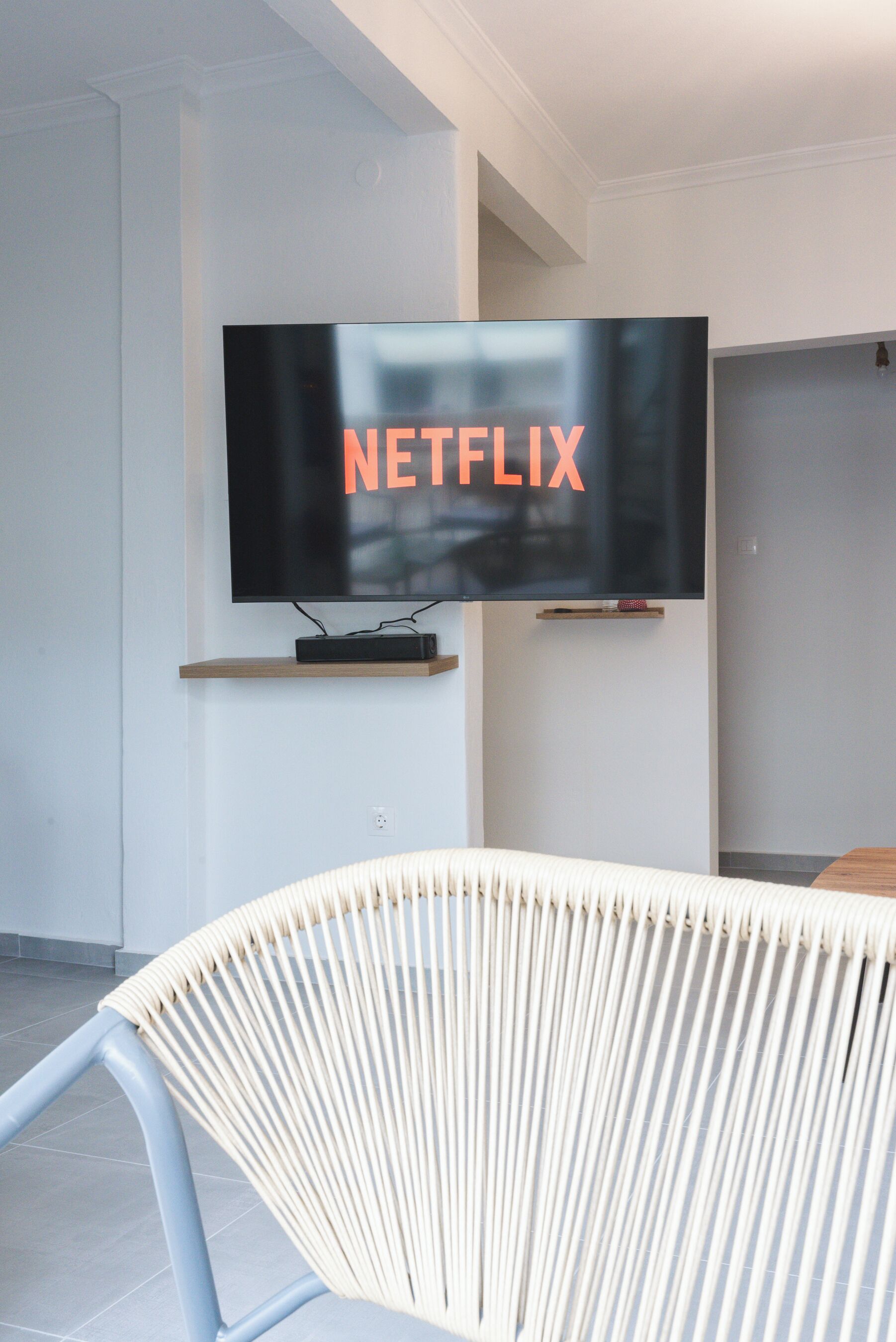 En moderne stue med et ophængt fjernsyn, der viser Netflix-logoet, en træhylde nedenunder og en hvid, vævet stol.