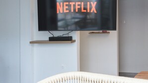 Ein modernes Wohnzimmer mit einem an der Wand befestigten Fernseher, der das Netflix-Logo zeigt, einem Holzregal darunter und einem weißen gewobenen Sessel.