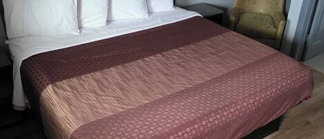 A neatly made bed with a patterned bedspread, a wooden floor, a chair, and a wall-mounted light fixture.