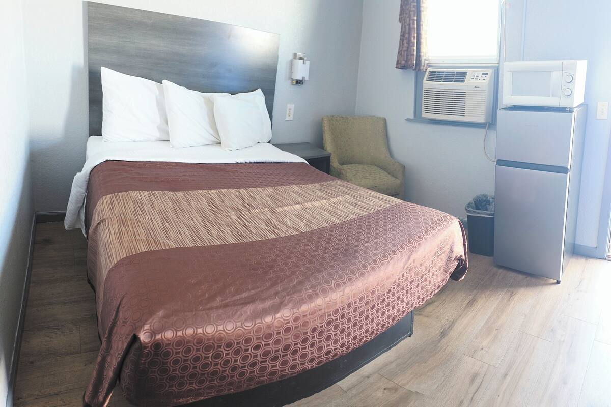 A hotel room with a bed, a chair, an air conditioning unit, and a refrigerator.
