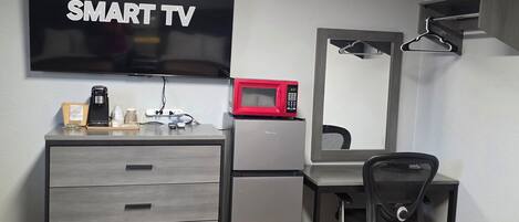 A modern room with a flat-screen TV labeled 'SMART TV,' a compact refrigerator, a microwave, a desk with a chair, and a large mirror.