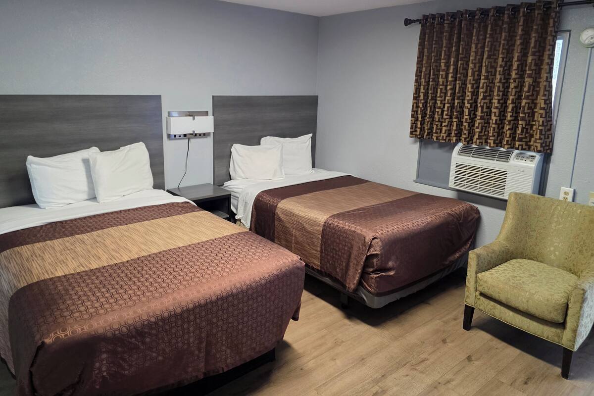 A hotel room with two beds, a chair, a wall-mounted air conditioner, and a power outlet.