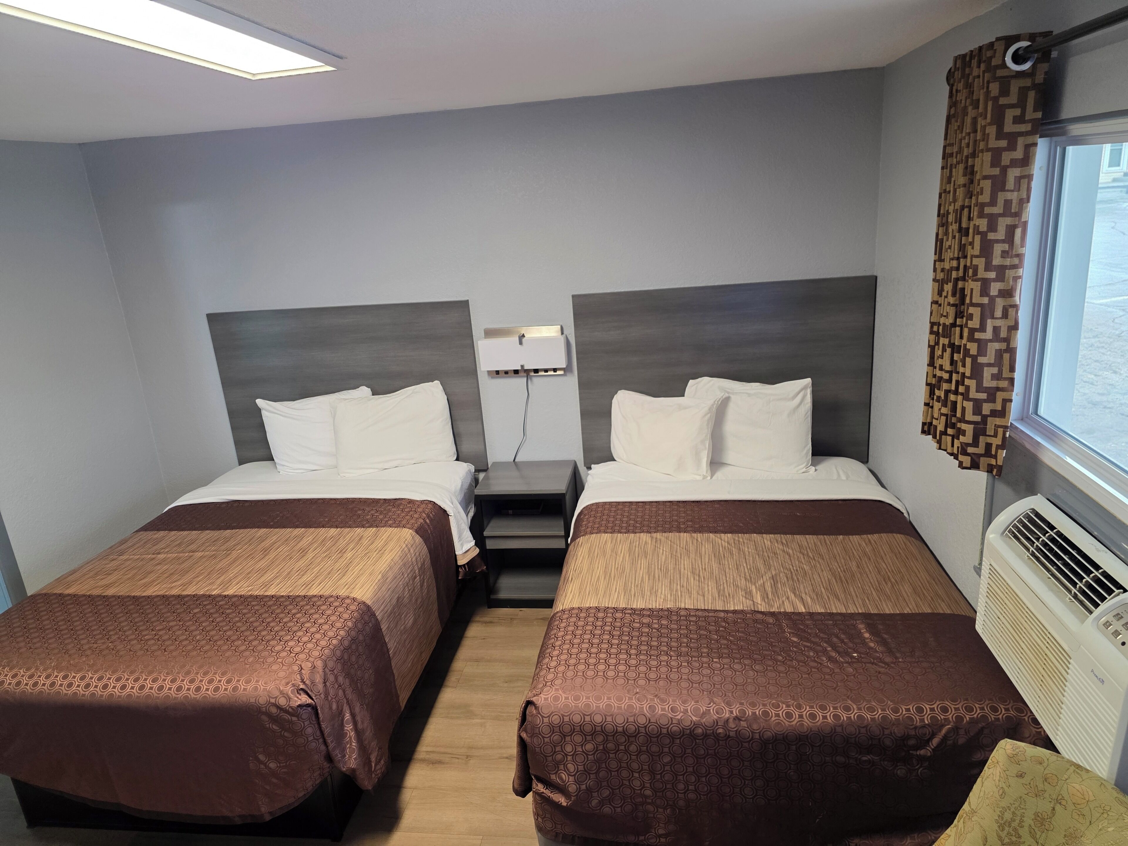 Habitación de hotel con dos camas, una mesita de noche, un ventanal con cortinas estampadas y un aire acondicionado de pared.