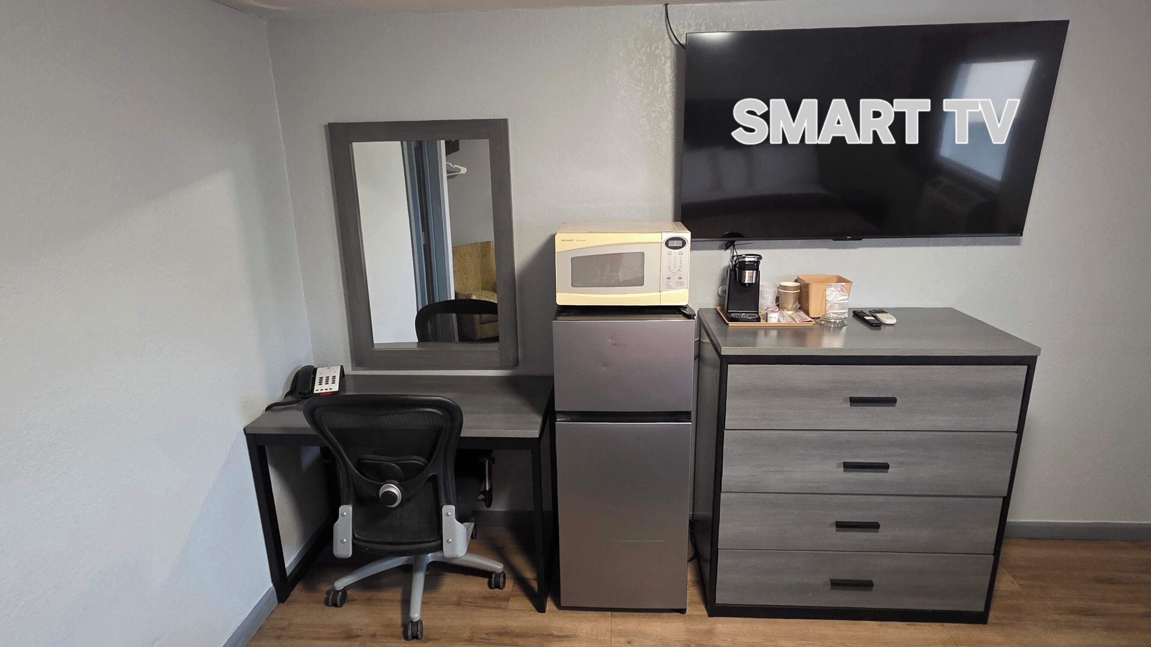 A modern hotel room with a desk, a chair, a microwave, a refrigerator, a coffee maker, and a flat-screen TV labeled 'SMART TV'.