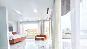 A modern hotel room with a bed, a sofa, a dining table, and a view of the city.