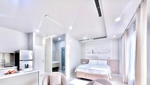 A modern hotel room with a kitchen area, a bed with a large window, and two armchairs.
