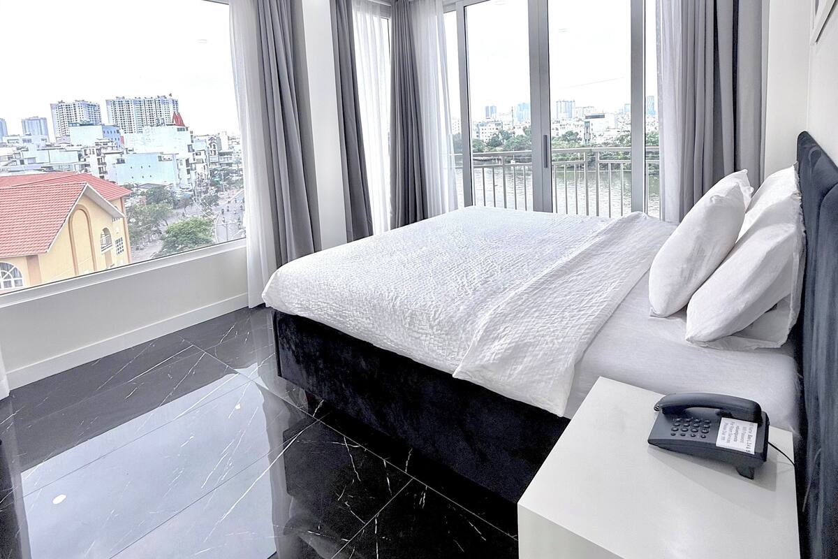 A modern hotel room with a large bed, a bedside phone, and a city view through large windows.