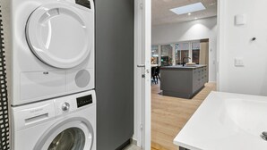 A modern laundry room with stacked washers and dryers, a kitchen area with a central island, and a bathroom with a white sink and mirror.