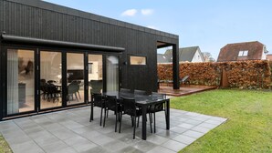 A modern house with a patio area featuring a table and chairs, a sliding glass door leading to an interior space, and a well-maintained garden with a hedge.