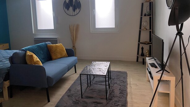 A living room with a blue sofa, a small table, a rug, a TV on a tripod stand, and a bookshelf. - Cosy studio in Évreux - Ideal! (Évreux)