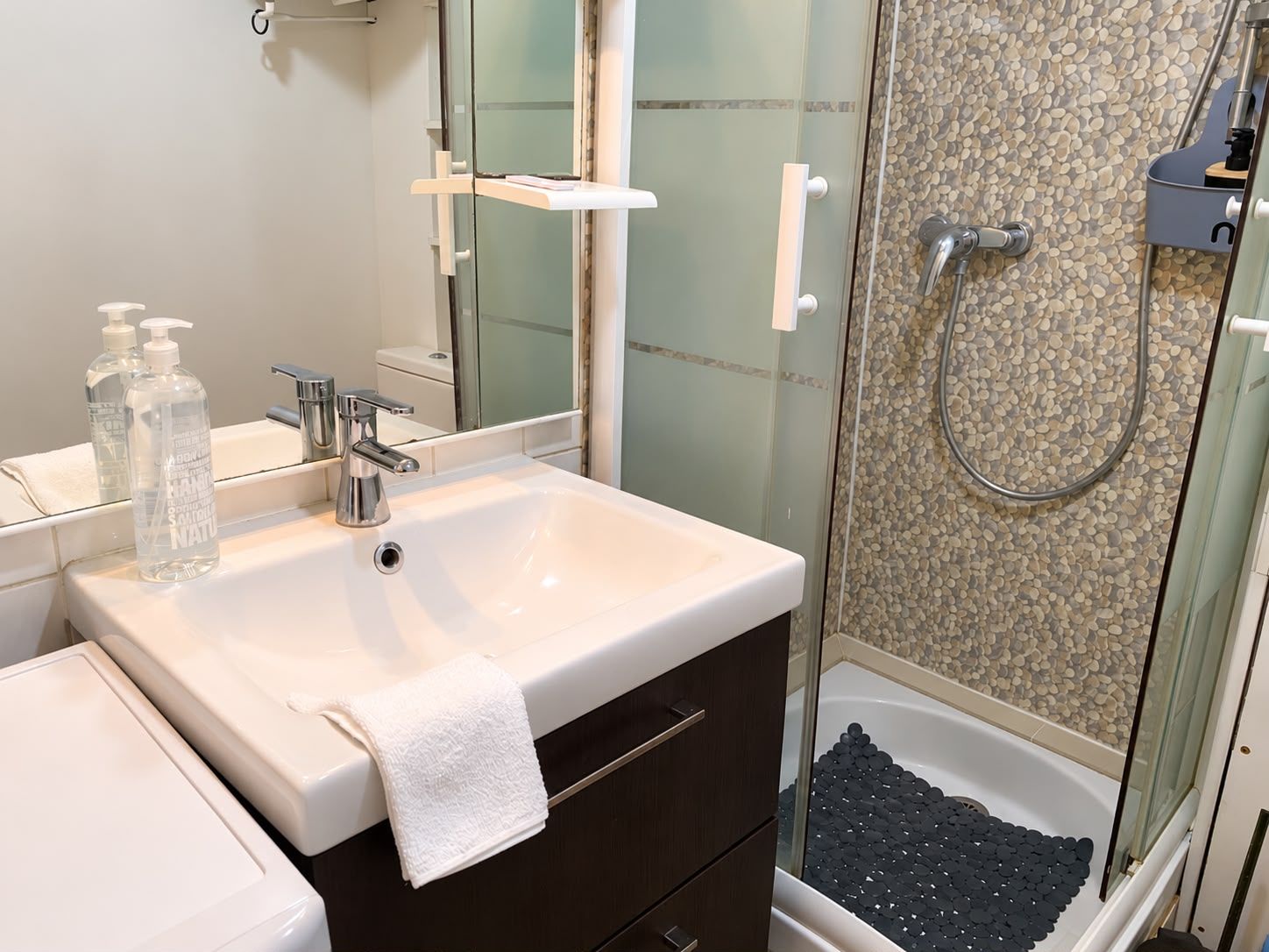 A bathroom with a white sink, a mirror, a towel, and a shower area with a pebble wall.