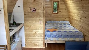 Une cabane chaleureuse avec un intérieur en bois, un lit recouvert d’une courtepointe, une petite fenêtre et un coin cuisine.