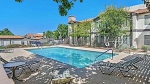 A swimming pool with lounge chairs and a table, surrounded by a fence and trees. - Contemporary luxurious Condo with Mountain View’s in Henderson . WiFi, Roku TV.  (Henderson)