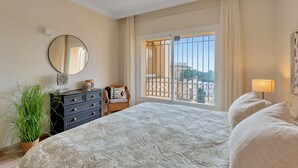 A bedroom with a large bed, a dresser, a chair, a potted plant, a round mirror, and a window with a view of buildings. - Charming Villa With Terrace (Marbella)