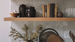 Una cocina con estantes de madera que sostienen utensilios de cocina, un cuadro enmarco y un jarrón con plantas secas.