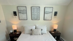 A neatly made bed with a white comforter and pillows, flanked by two bedside tables with lamps, and three framed artworks on the wall above. - The Innwoods - Studio Apartment 1 (Tannersville)