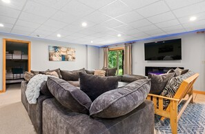 Een moderne woonkamer met een hoekbank, een houten bankje, een flatscreen televisie en een groot raam met gordijnen.
