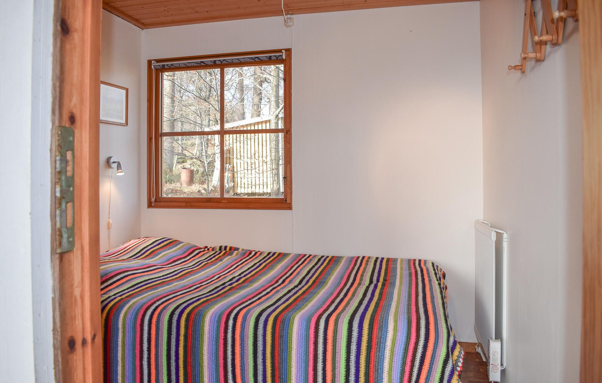 Una habitación pequeña con una cama individual, una colcha a rayas de colores, una ventana con vistas a árboles y un radiador en la pared.