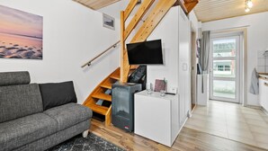 A modern living room with a grey sofa, a TV mounted on a wooden beam, a small white cabinet, and a staircase leading to an upper level. - 2 bedroom stunning home in Fanø (Fanø)