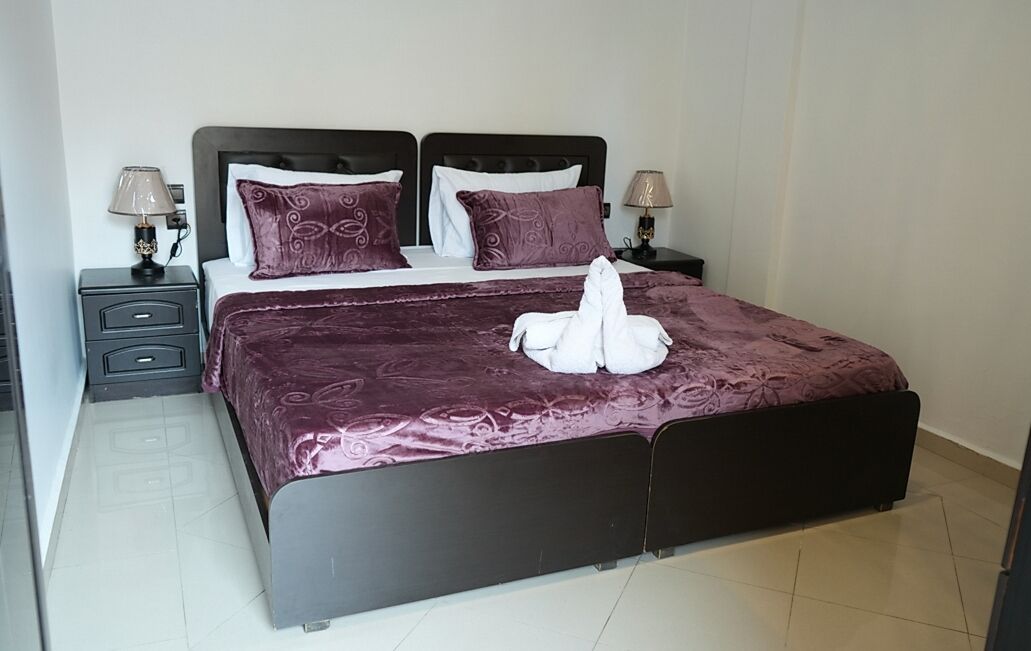 A neatly made bed with a purple bedspread and pillows, two bedside tables with lamps, and a folded white towel on the bed.