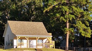 A small, single-story house with a porch, surrounded by a grassy field and a large tree. - Cozy Cabin by the Lake (Tyler)