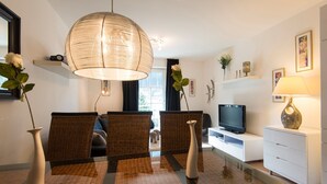 Un salon moderne avec une table à manger en verre, des chaises en osier, un grand luminaire tressé, un téléviseur à écran plat et un meuble de rangement blanc.