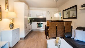 Une cuisine moderne avec des meubles blancs, un coin repas avec des meubles en osier et un salon doté d’un canapé en cuir noir.