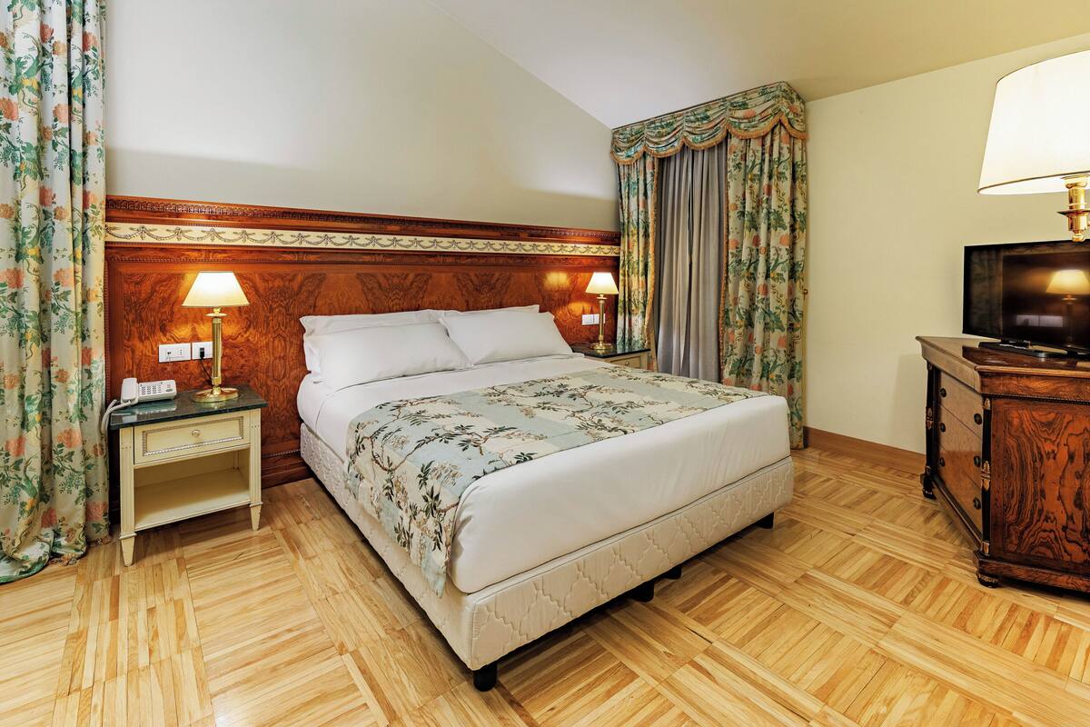 Camera d'albergo con una testiera in legno, un letto con biancheria da letto floreale, un comodino con una lampada, una cassettiera con televisore e tende a fiori.