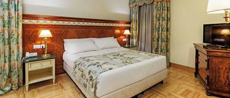 A hotel room with a wooden headboard, a bed with floral bedding, a nightstand with a lamp, a dresser with a TV, and floral curtains.