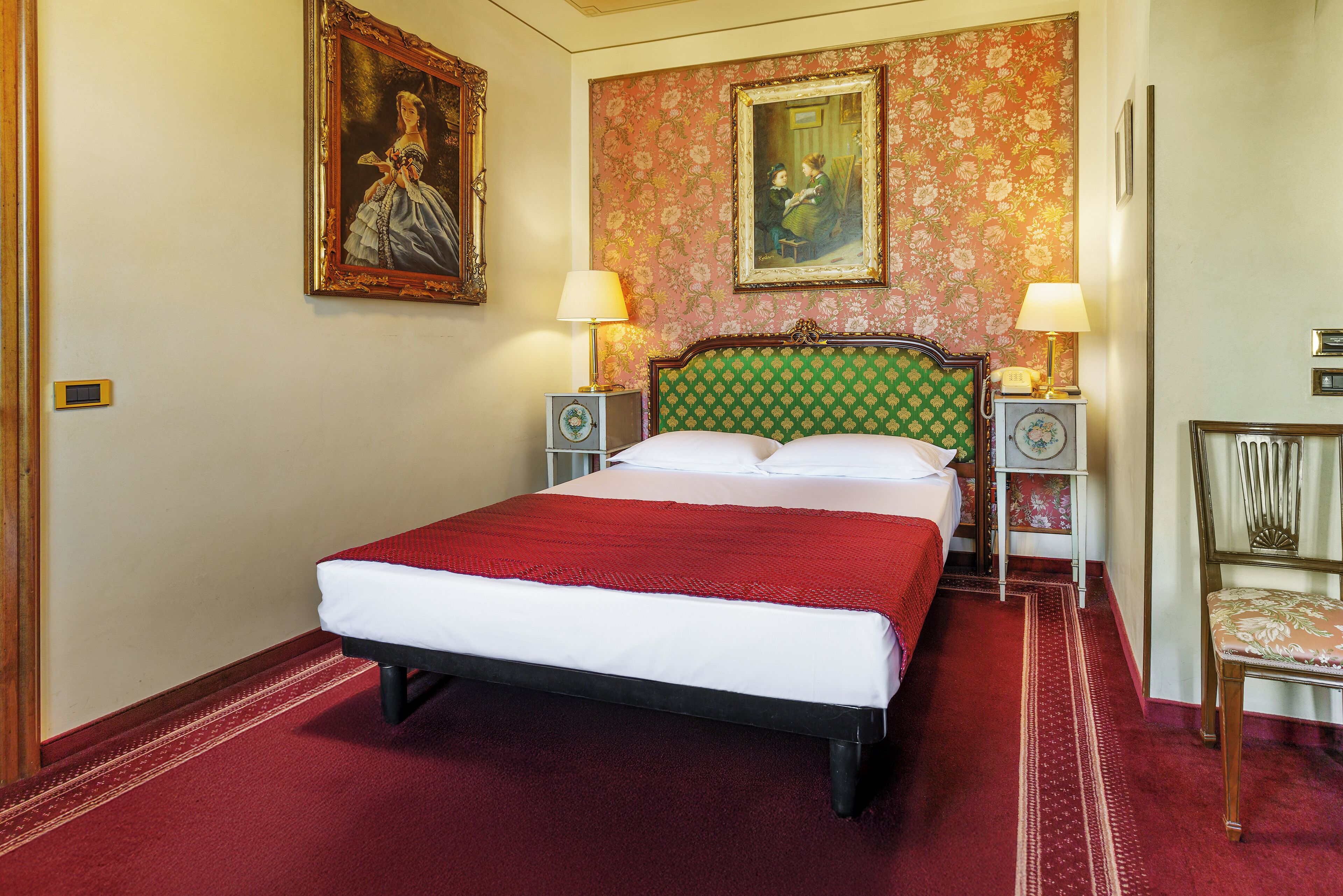 A hotel room with a large bed, two bedside tables with lamps, a chair, and floral wallpaper.