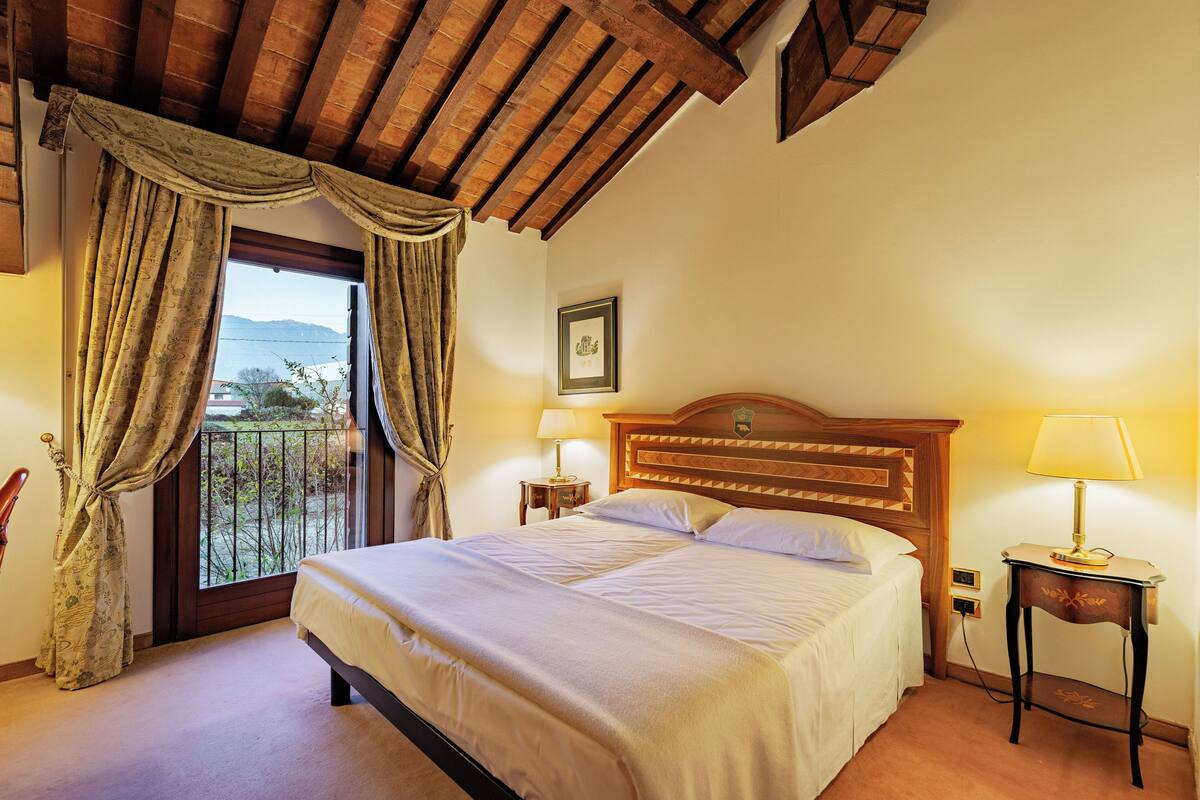 Una camera d'albergo con un letto, un comodino con una lampada, un quadro appeso al muro e una finestra con tende.