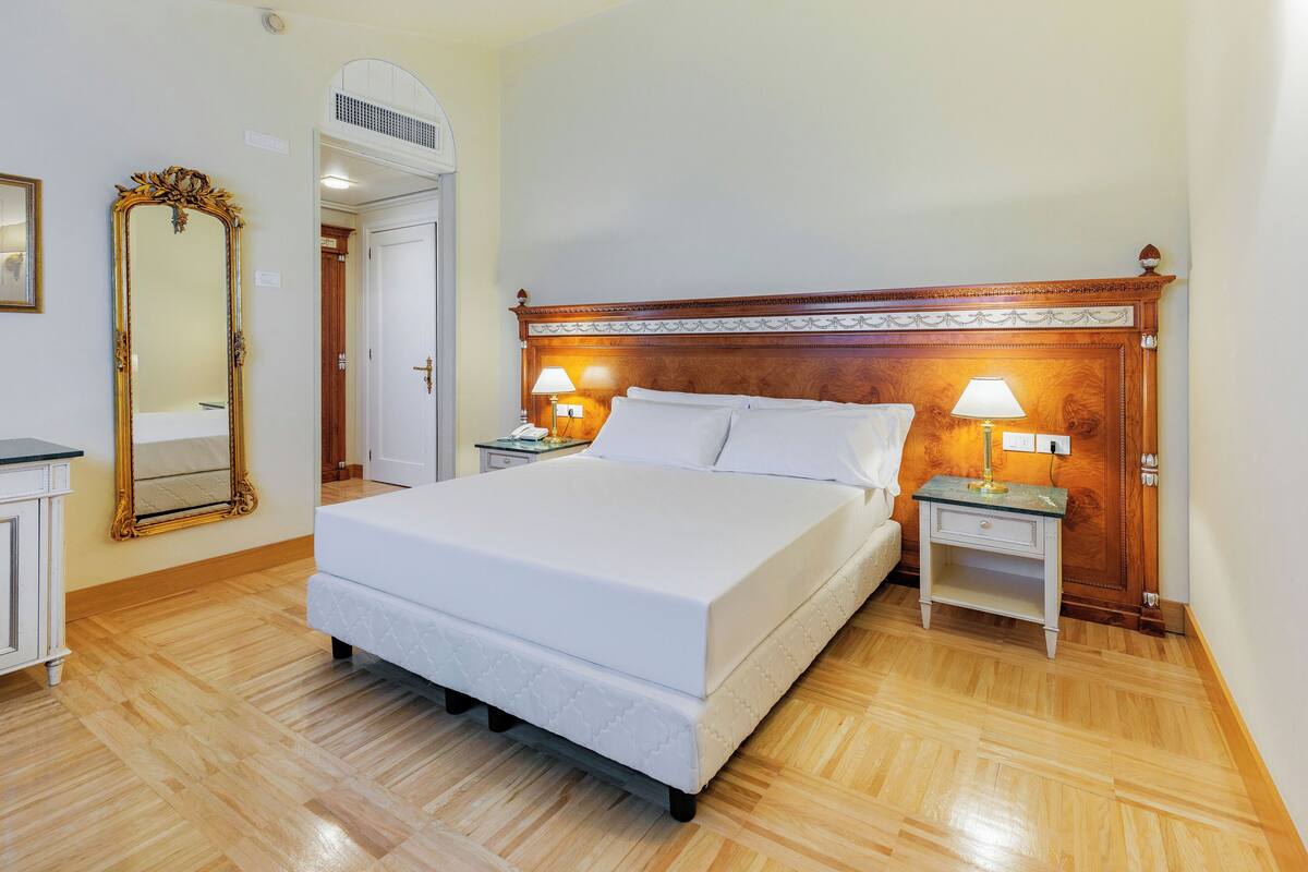 Una camera da letto ordinata con un letto grande, due comodini con lampade, una testiera in legno e uno specchio a figura intera, con pavimento in legno.
