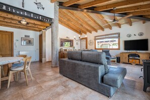 A spacious living area with a grey sofa, wooden beams, a dining table with chairs, a TV, and a fireplace.