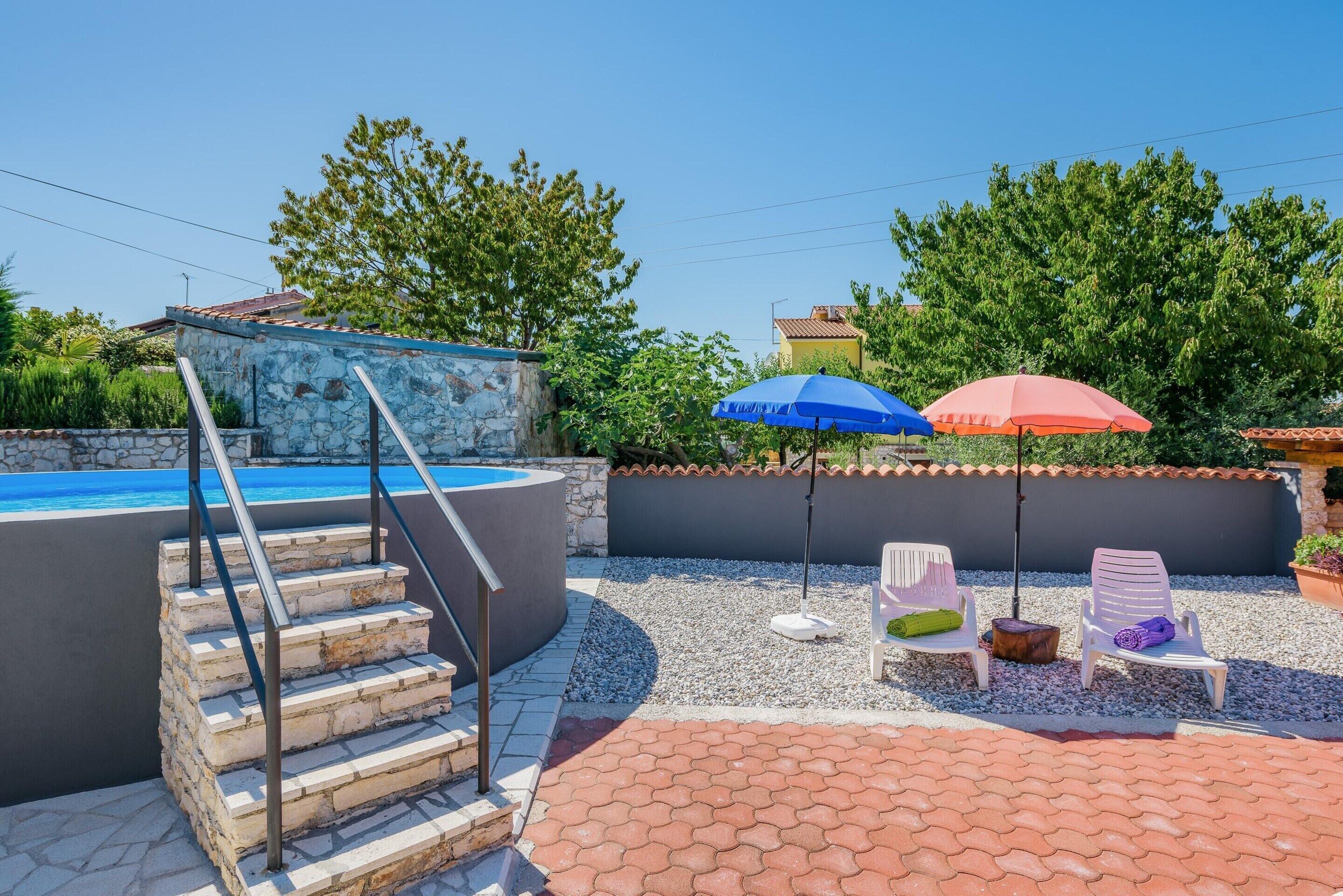 A swimming pool with steps leading down, surrounded by a pebble-covered patio with two chairs and two umbrellas.