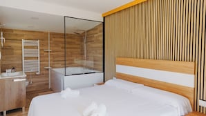 A modern hotel room with a wooden floor, a large bed with white linens, a glass-enclosed shower area, a wooden headboard, and a wooden vanity with a sink and mirror. - WELCS DUPLEX 192 PDA cerca del mar (Platja d´Aro)
