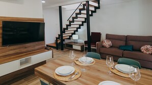 A modern living room with a wooden dining table set for two, a brown sofa, a TV mounted on a wooden wall, and a staircase. - WELCS DUPLEX 192 PDA cerca del mar (Platja d´Aro)