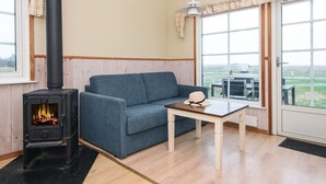 A cozy room with a blue sofa, a small wooden table, a black stove with visible flames, and large windows offering a view of the outdoors. - 2 Person Holiday Park Home in Romo (Rømø)