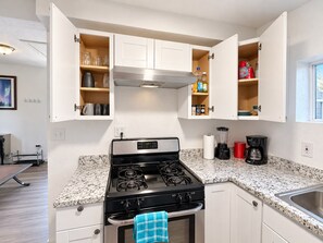 Una cocina con estufa a gas, horno empotrado y pileta de acero inoxidable. Se observan gabinetes con las puertas abiertas que muestran utensilios de cocina y especias.