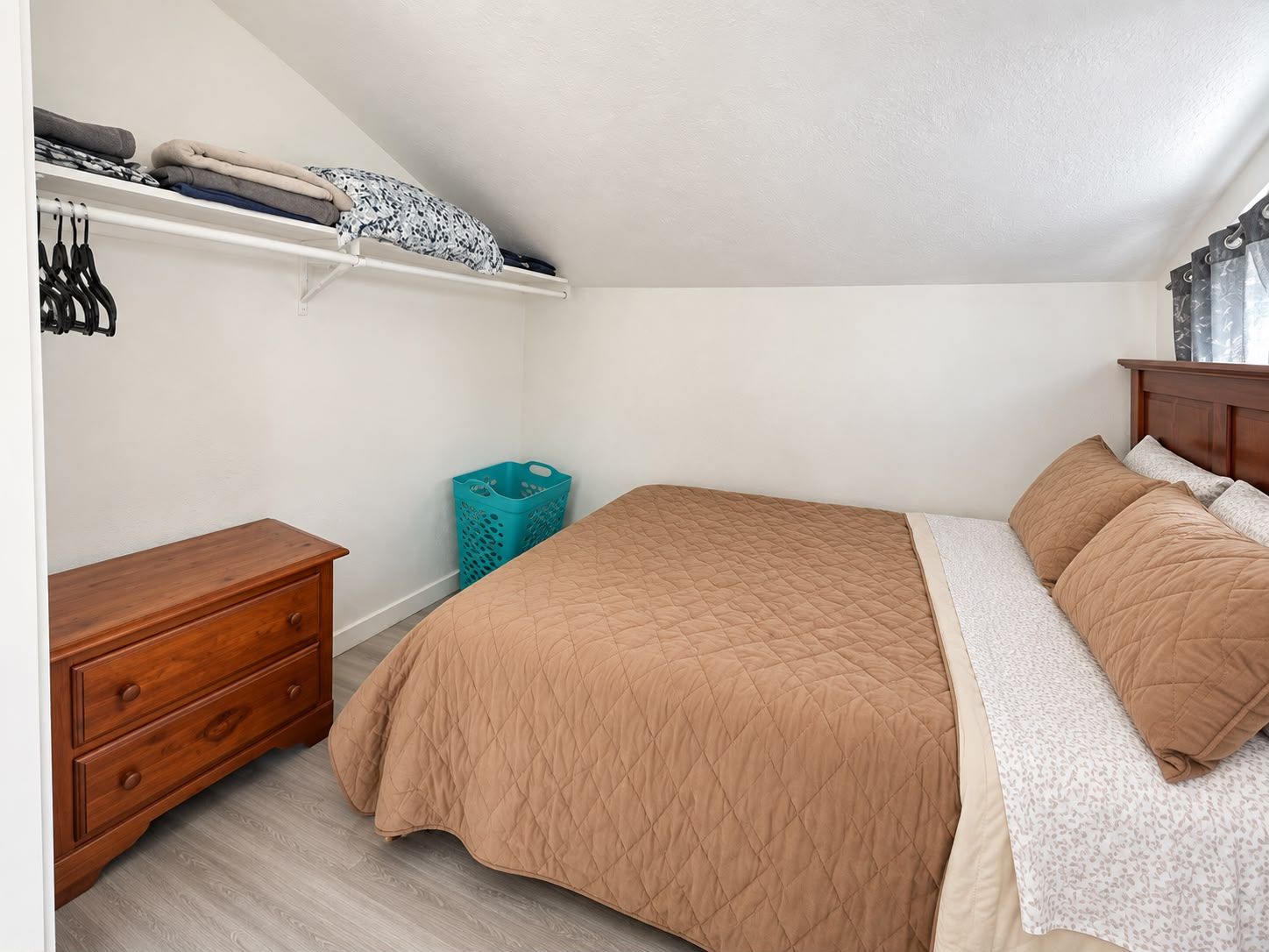 Een slaapkamer met een bed, een houten nachtkastje, een dressoir, een plank met opgevouwen kleding en een wasmand.