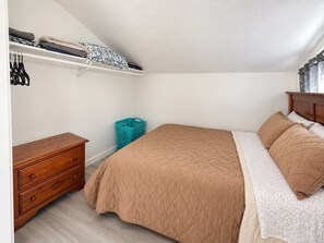 Een slaapkamer met een bed, een houten nachtkastje, een dressoir, een plank met opgevouwen kleding en een wasmand.