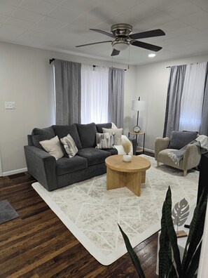 Een woonkamer met een grijze bank, een ronde houten salontafel, een beige fauteuil en een plafondventilator.