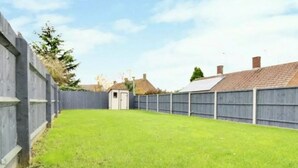 Une cour arrière clôturée avec une pelouse verdoyante, un petit abri de jardin et une maison avec un toit en tuiles en arrière-plan.