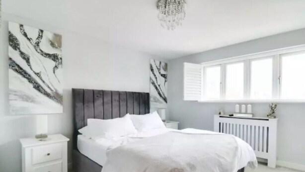 Une chambre à coucher avec un lit, des tables de chevet blanches, un radiateur et des œuvres d’art accrochées aux murs.