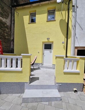 A yellow building with a white door, two windows, and a small porch with a chair.