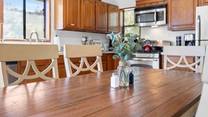 Una mesa de comedor de madera con sillas, un florero con flores y electrodomésticos de cocina al fondo.