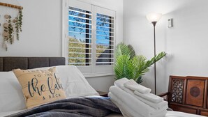 Una cama bien hecha con una almohada, una lámpara de noche, una pila de toallas y una ventana con persianas.
