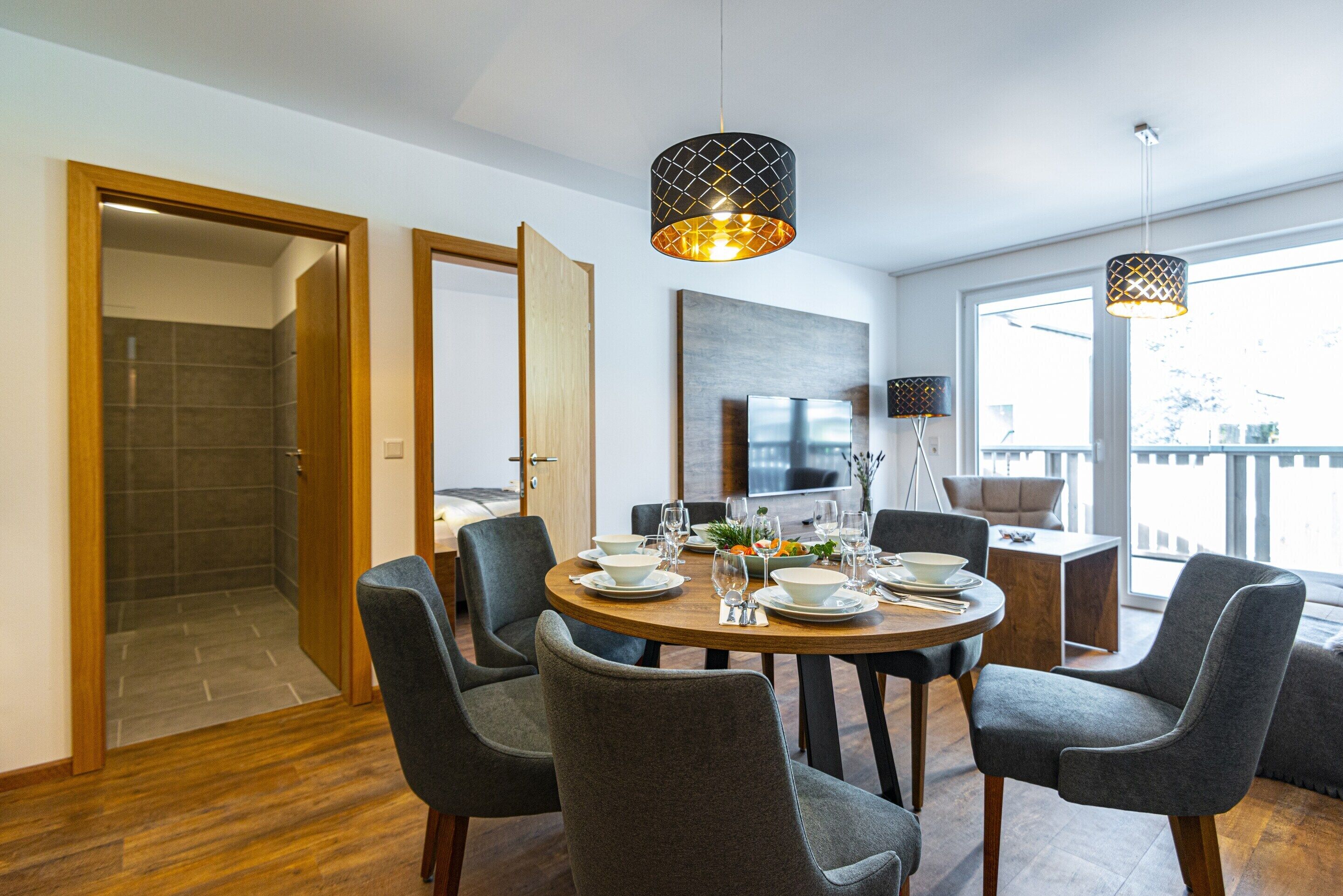 A dining area with a round table set for four, surrounded by grey upholstered chairs. There are two pendant lights above the table, and a flat-screen TV mounted on the wall.